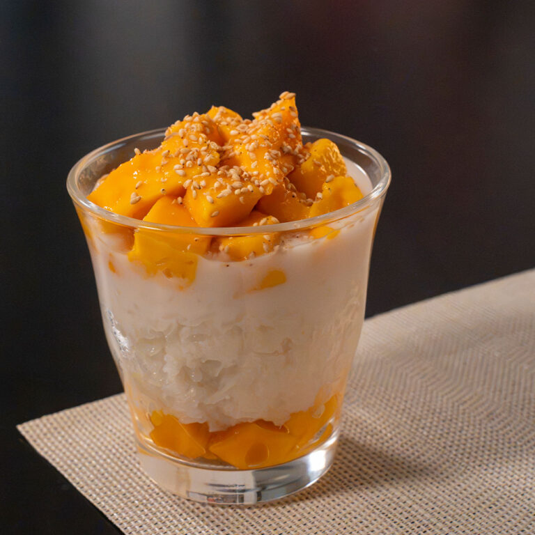 A single-serving mango sticky rice dessert layered in a clear glass with creamy coconut sticky rice, sweet ripe mango chunks, and a crunchy toasted topping.
