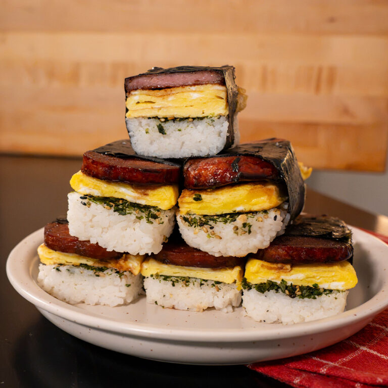 Stacked spam musubi with rice, egg, and nori on a white plate.