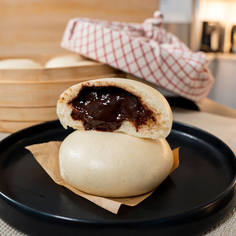 Choco-Pao chocolate steamed buns on a black plate, with the top bun torn open to show melted chocolate ganache filling and a bamboo steamer in the background
